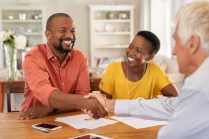 "A couple shaking hands with a man across the table, likely in a business or formal meeting setting, with papers and phones visible on the table."