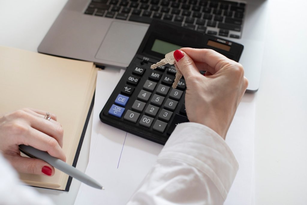 "A person holding a set of keys while looking at a calculator and a laptop, with a notebook and pen beside them, possibly calculating expenses or making financial decisions."